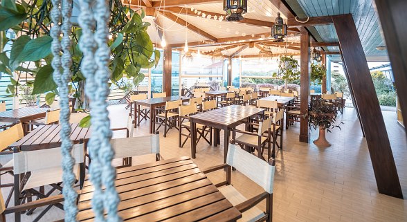  Restaurant with indoor and covered outdoor area with many wooden tables and chairs, decorated with hanging plants and warm lamps.