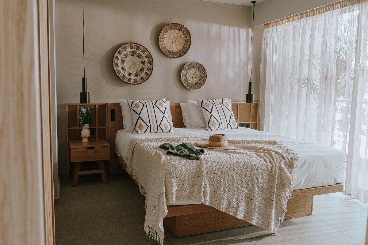 Bright bedroom with two beds, woven wall plates, bedspread and sun hat on the bed, light through curtain.